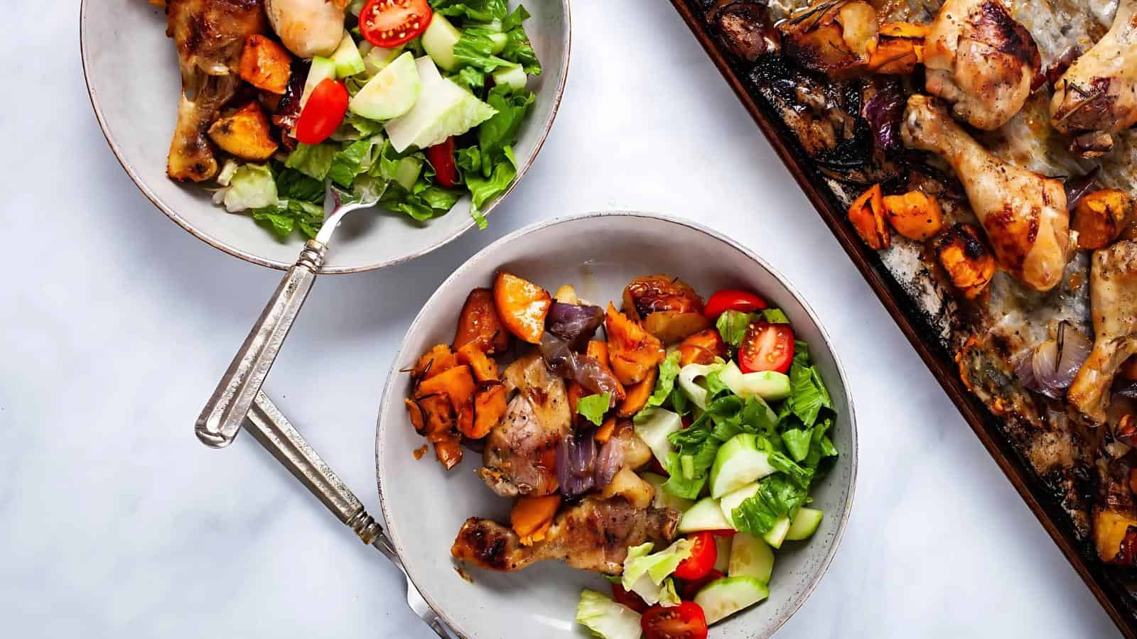 Two plates with chicken drumsticks, roasted vegetables, lettuce, cucumbers, and tomatoes. A baking tray with more chicken and vegetables is visible next to the plates.