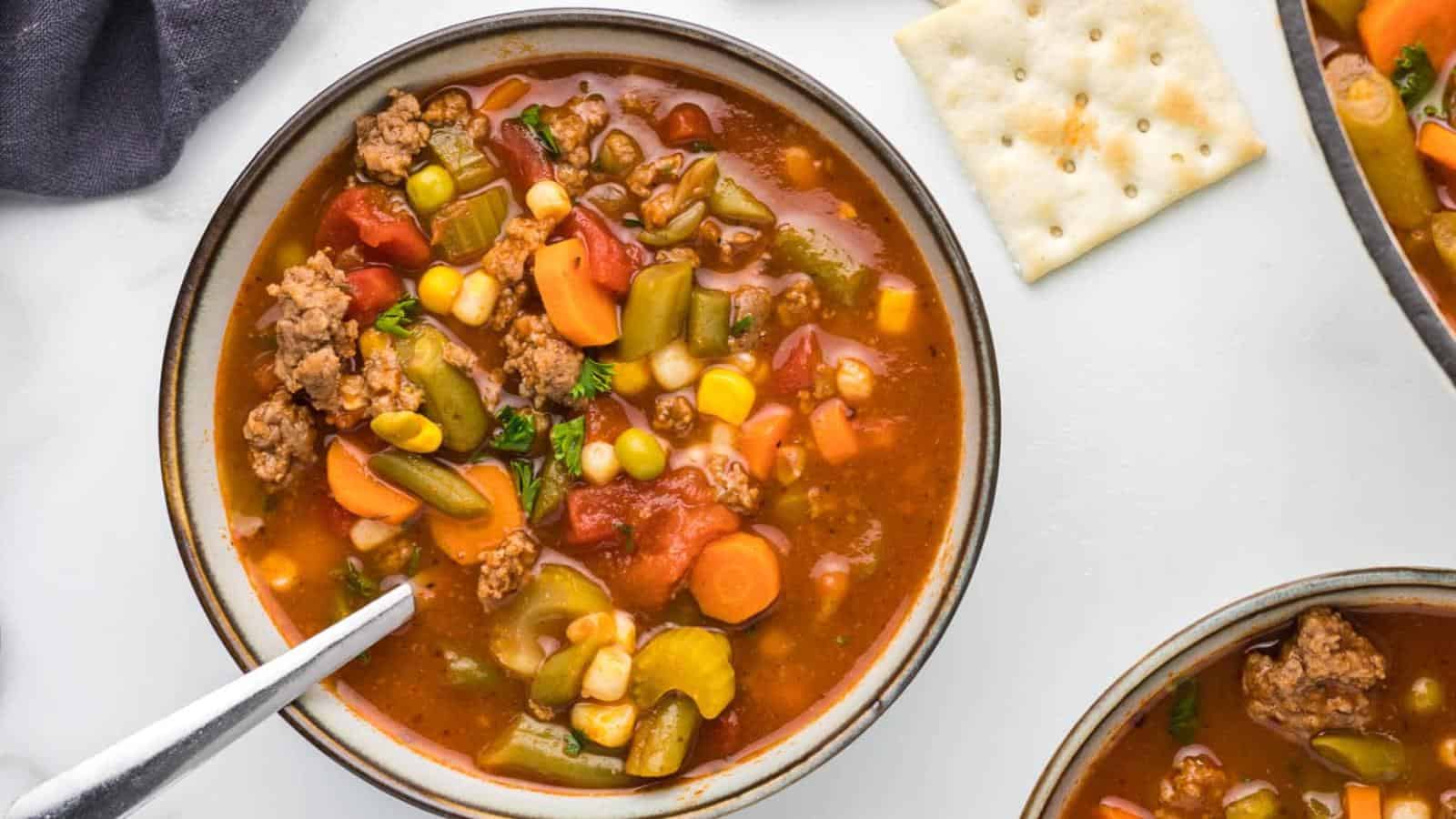 Bowl of vegetable beef soup with visible carrots, corn, green beans, and tomatoes, next to a cracker on a white surface.