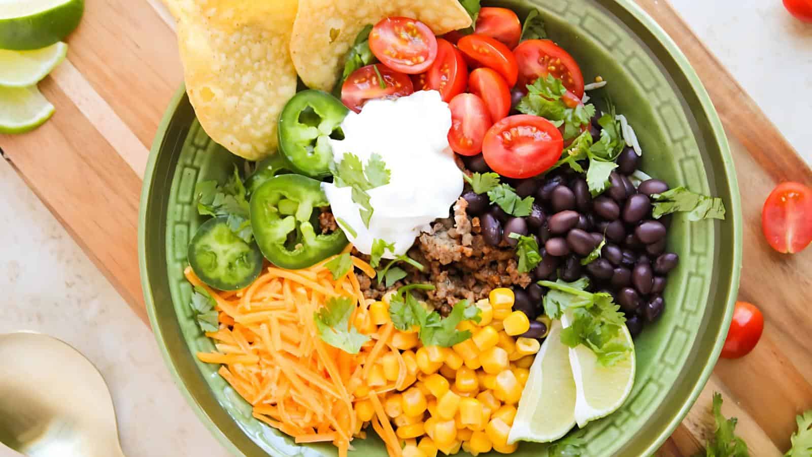 A green bowl filled with cherry tomatoes, black beans, corn, shredded cheese, ground meat, jalapeño slices, cilantro, tortilla chips, lime wedges, and a dollop of sour cream.
