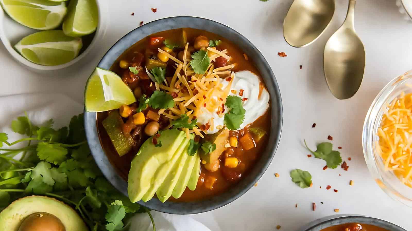 A bowl of vegetable chili garnished with avocado slices, sour cream, shredded cheese, and cilantro, accompanied by lime wedges. Nearby are spoons and fresh cilantro.