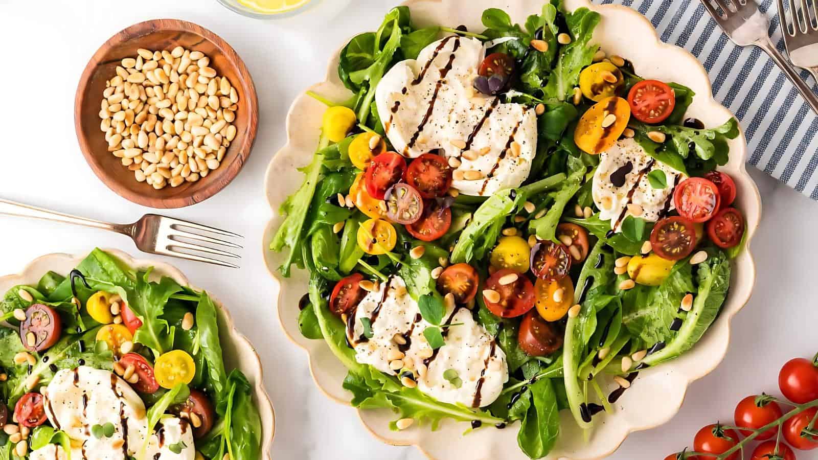 Caprese salad with mozzarella, cherry tomatoes, arugula, and pine nuts on a white plate, drizzled with balsamic glaze, with a side of pine nuts and a fork nearby.