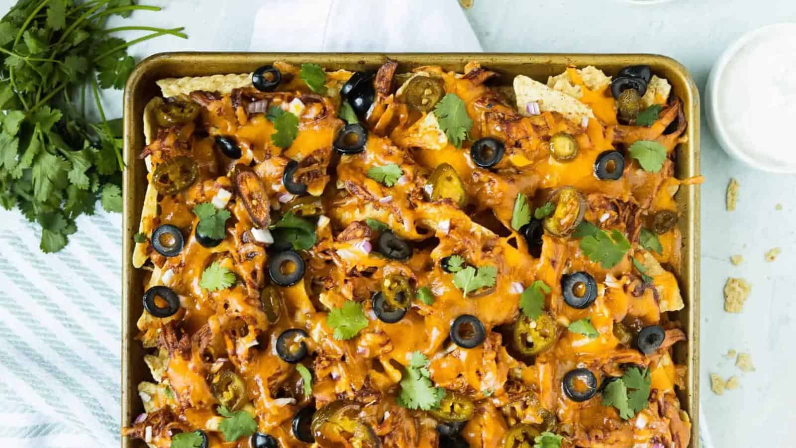 A baking tray filled with nachos topped with melted cheese, black olives, jalapeños, cilantro, and shredded chicken. A bunch of fresh cilantro is placed nearby.
