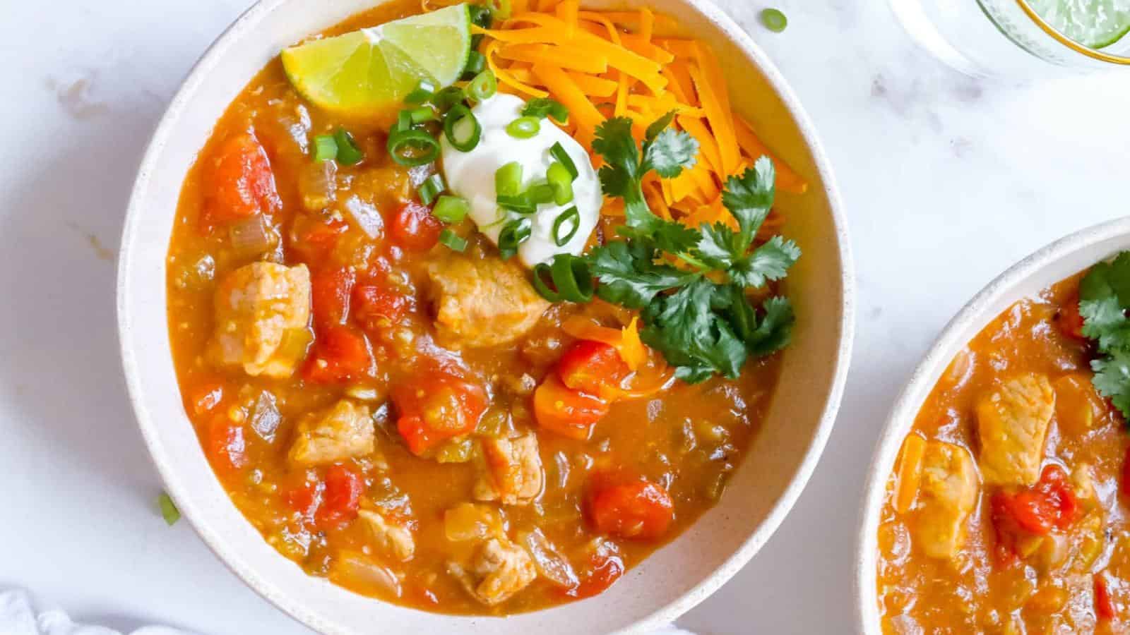 A bowl of chili with chunks of meat, tomatoes, and red peppers, topped with shredded cheese, sour cream, sliced green onions, cilantro, and a lime wedge.