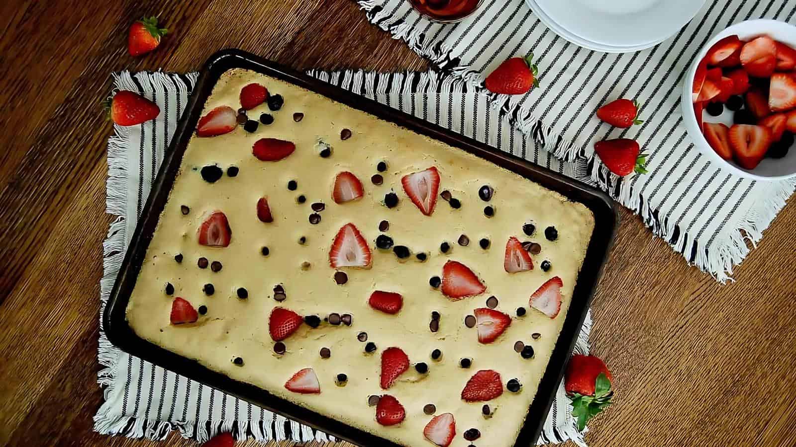 A rectangular cake on a striped cloth, topped with sliced strawberries and chocolate chips, surrounded by scattered strawberries and a bowl of cut berries.