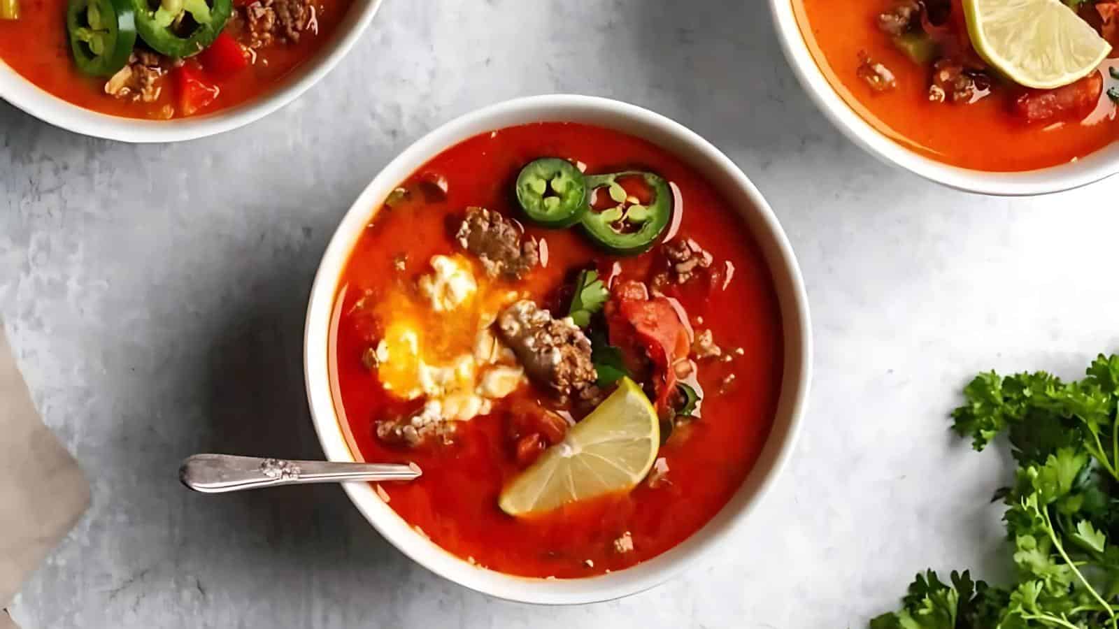 Bowl of red soup with ground meat, sliced jalapeños, lemon slice, and a spoon. Other bowls and cilantro are partially visible around it.