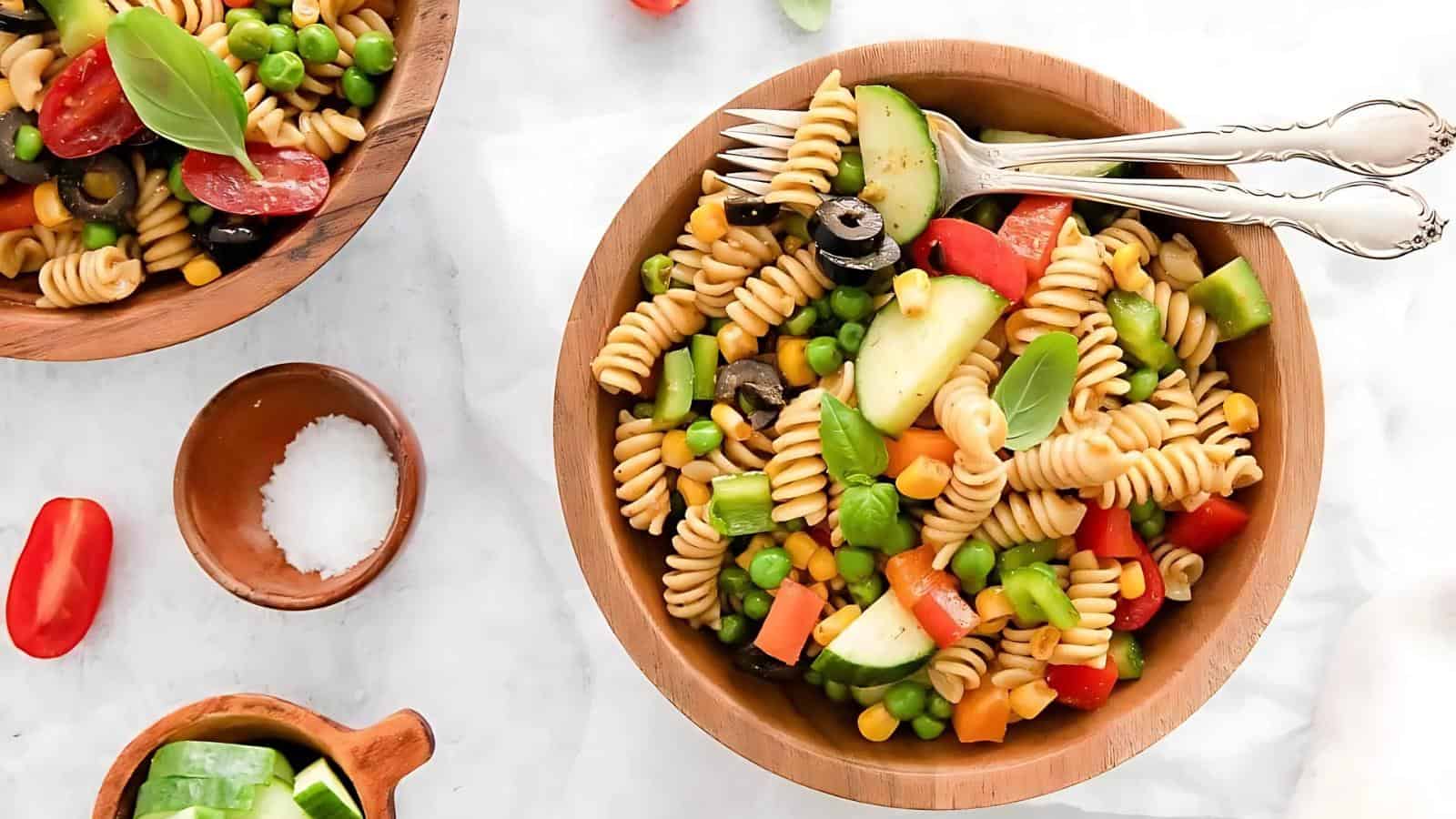 A wooden bowl of pasta salad with rotini, black olives, corn, peas, cucumber, bell peppers, and tomato, garnished with basil. Forks rest on the bowl's rim.