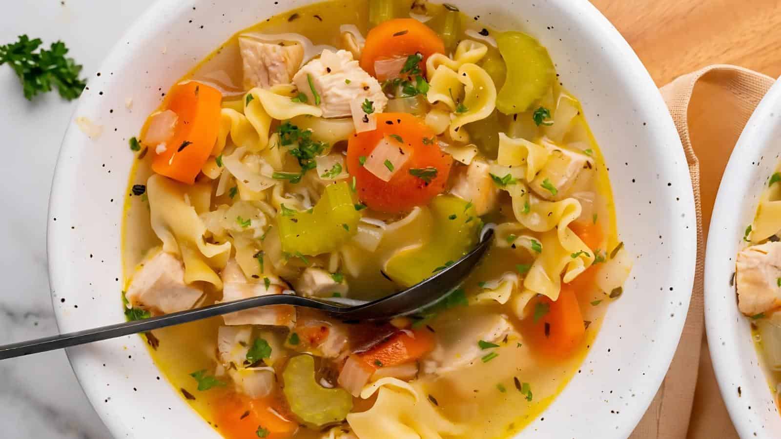 A bowl of chicken noodle soup with chunks of chicken, carrots, celery, egg noodles, and parsley, with a spoon resting inside.