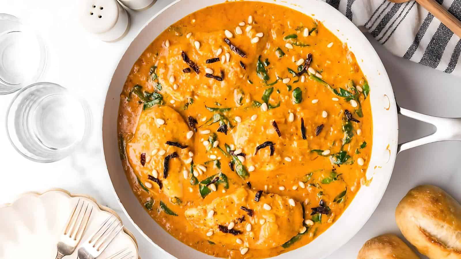 A skillet contains chicken in a creamy orange sauce, garnished with pine nuts and herbs. Two glasses, a striped cloth, and a salt and pepper shaker are nearby.