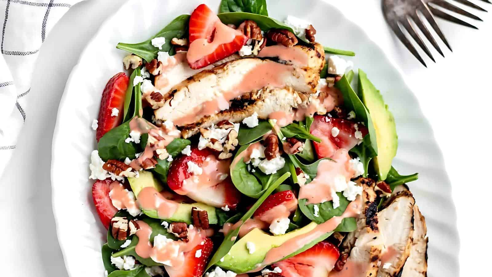 Salad with grilled chicken, strawberries, avocado, spinach, pecans, feta cheese, and pink dressing on a white plate.