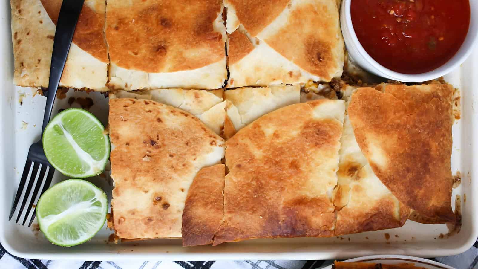 Sliced quesadillas on a tray with lime wedges and a bowl of red salsa.