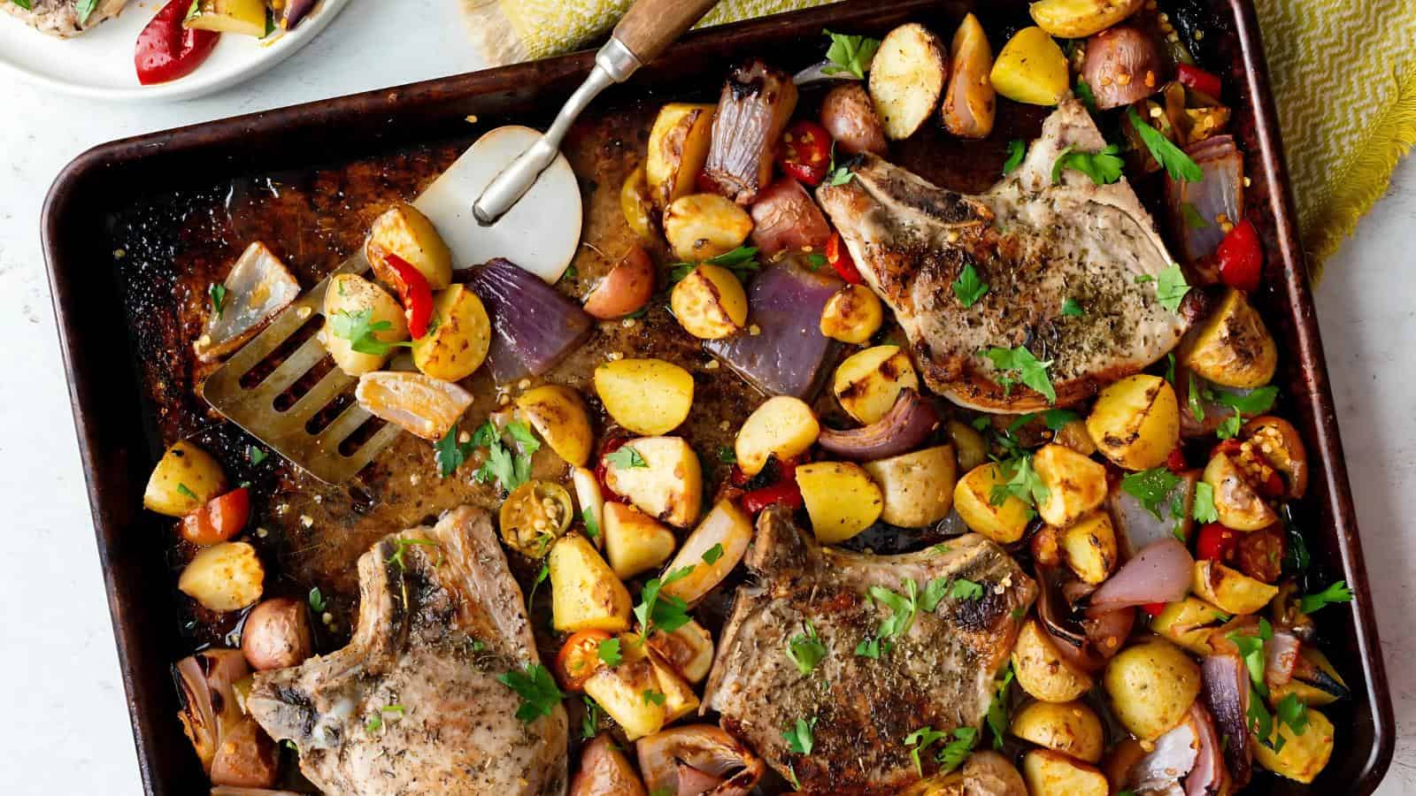 Sheet pan with pork chops, roasted potatoes, onions, cherry tomatoes, and herbs. A spatula and pizza cutter rest on the pan.