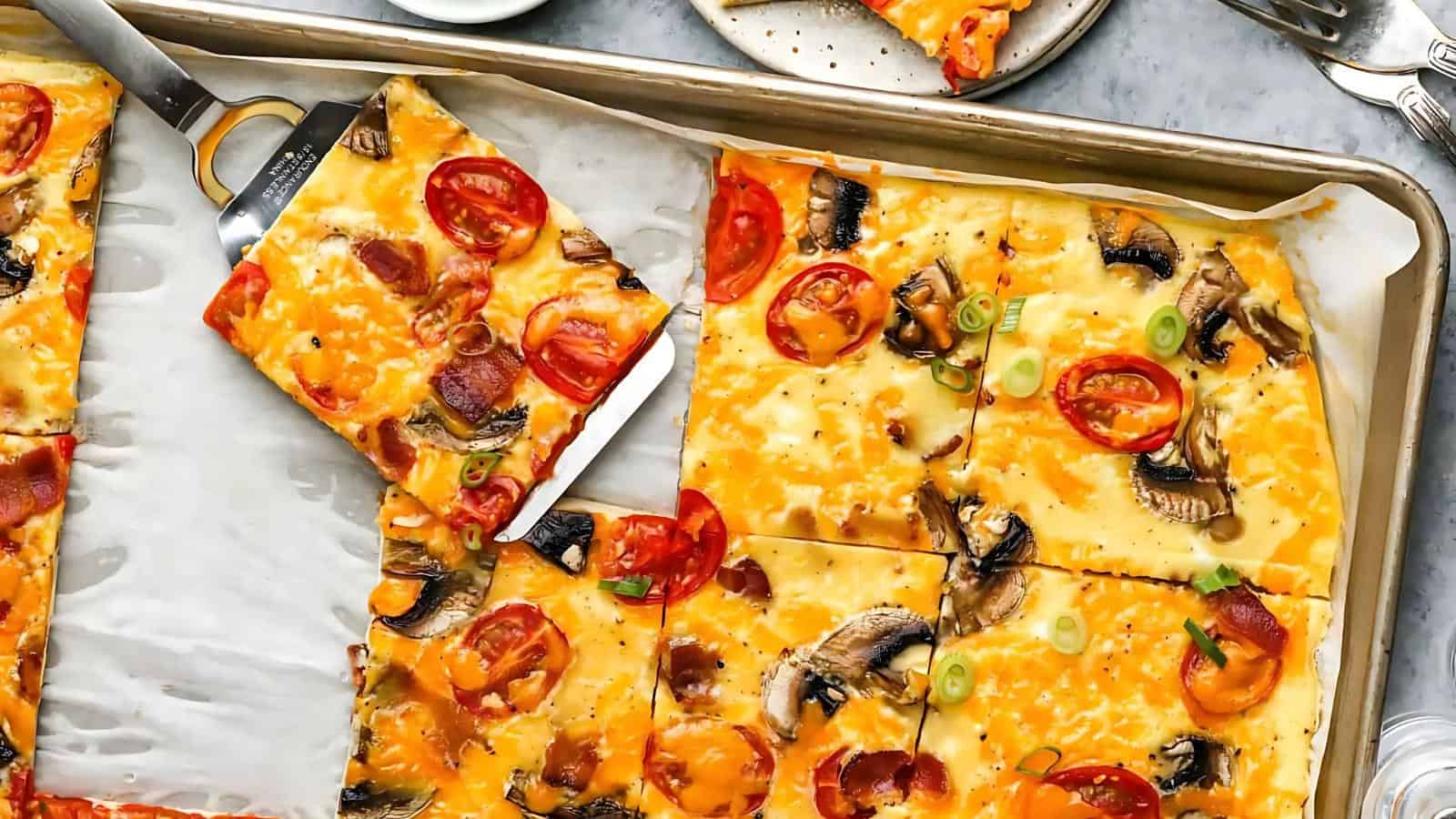 Sheet pan pizza with melted cheese, sliced tomatoes, mushrooms, pepperoni, and green onions. A spatula is lifting one square slice from the tray. Plates and utensils are nearby.