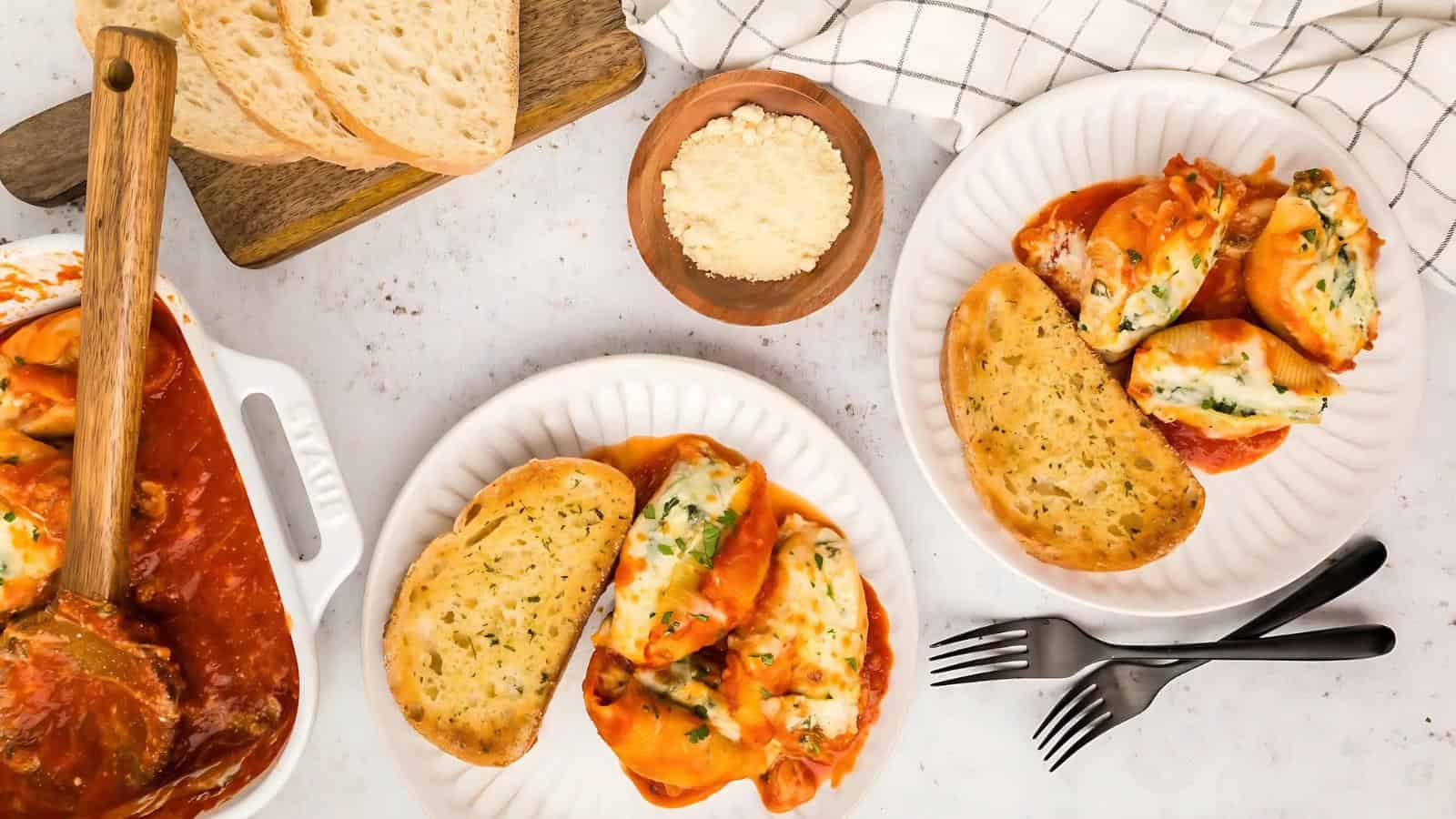 Two plates of stuffed pasta shells with tomato sauce and garlic bread. A casserole dish, parmesan cheese bowl, and sliced bread are nearby on a white surface.