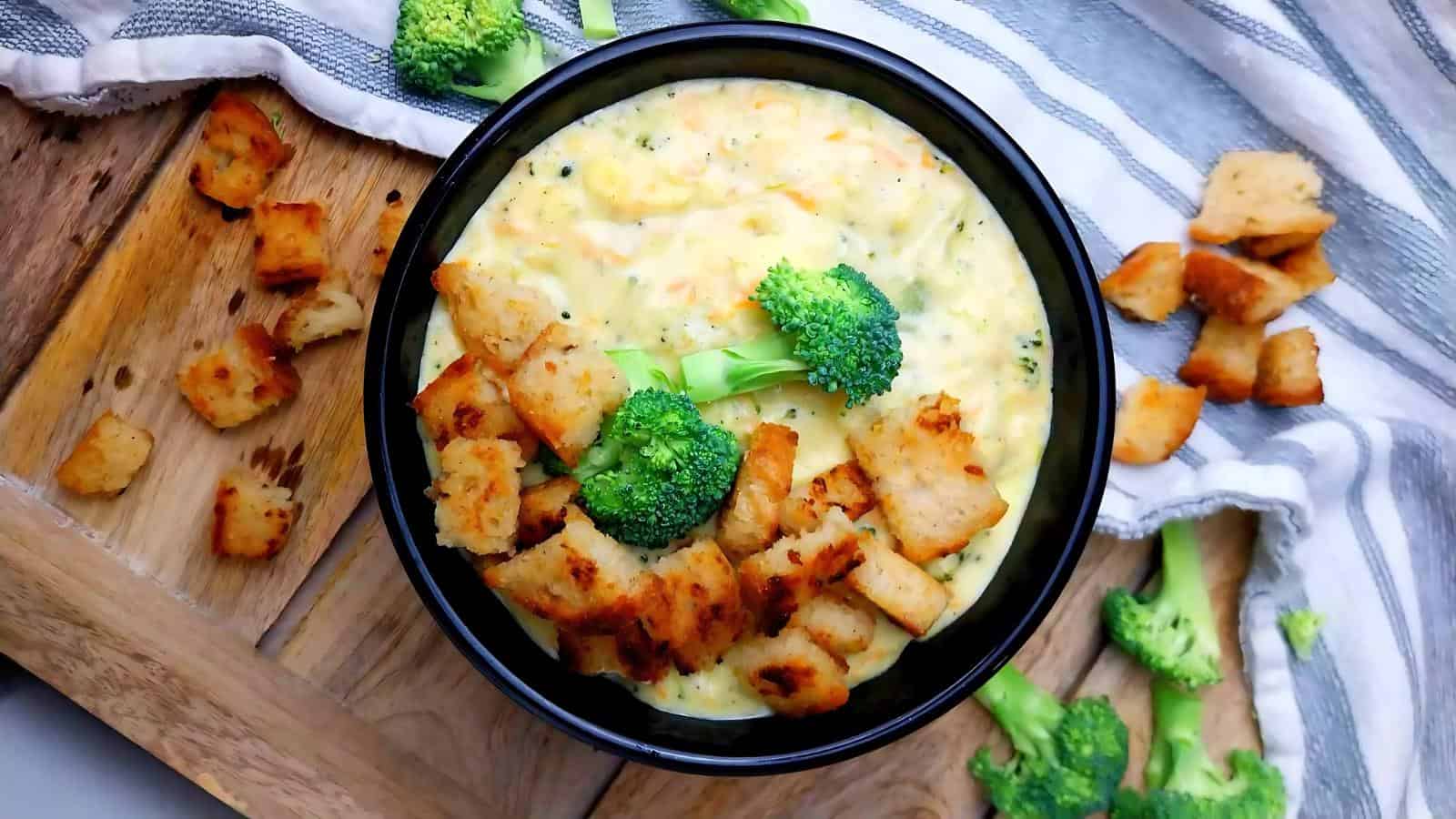 A bowl of broccoli cheese soup topped with croutons and broccoli florets, placed on a wooden surface with scattered croutons and a striped cloth.