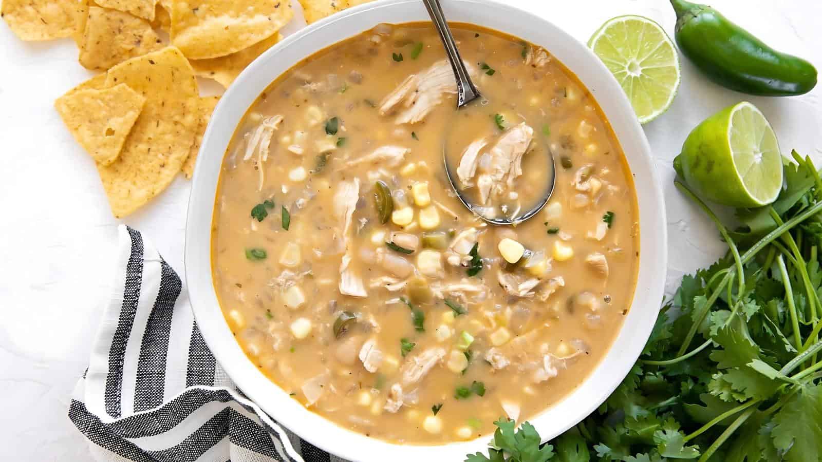 Bowl of creamy chicken soup with corn and herbs, garnished with cilantro and served with tortilla chips. Lime and jalapeño are on the side.