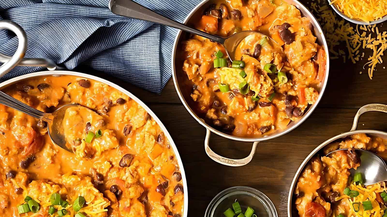 Bowls of creamy taco soup with ground meat, black beans, and vegetables on a wooden table. Topped with green onions and surrounded by shredded cheese and a blue cloth.