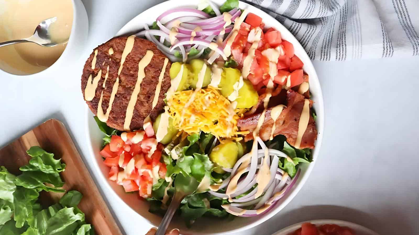 A salad in a bowl with lettuce, tomato, red onion, pickles, shredded cheese, burgers, bacon, and dressing drizzle. A bowl of dressing and a wooden board with lettuce on the side.