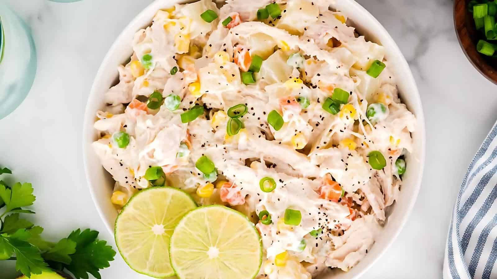 Bowl of creamy salad with shredded chicken, corn, peas, and diced vegetables, garnished with chopped green onions and lime slices.