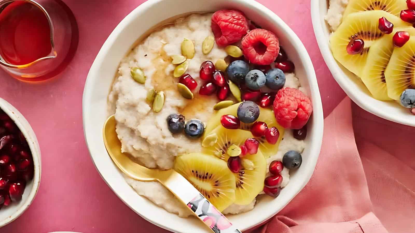 A bowl of oatmeal topped with sliced kiwi, raspberries, blueberries, pomegranate seeds, and pumpkin seeds, with a gold spoon on a pink background.