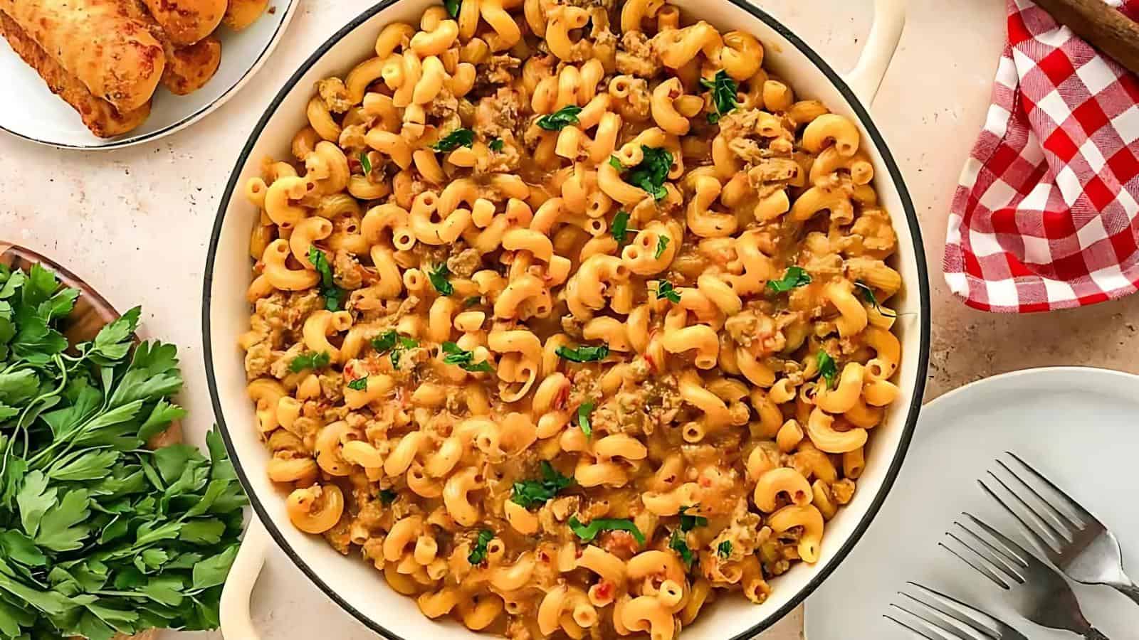 A pot of cooked macaroni with creamy sauce and ground meat, garnished with parsley. Nearby are a plate of bread and a checkered napkin with forks.