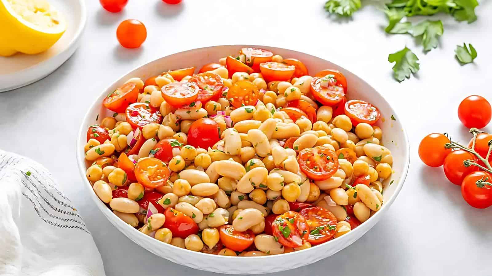 A bowl of bean salad with chickpeas, white beans, cherry tomatoes, red onion, and herbs. Chopped parsley and whole cherry tomatoes are scattered around on a light surface.
