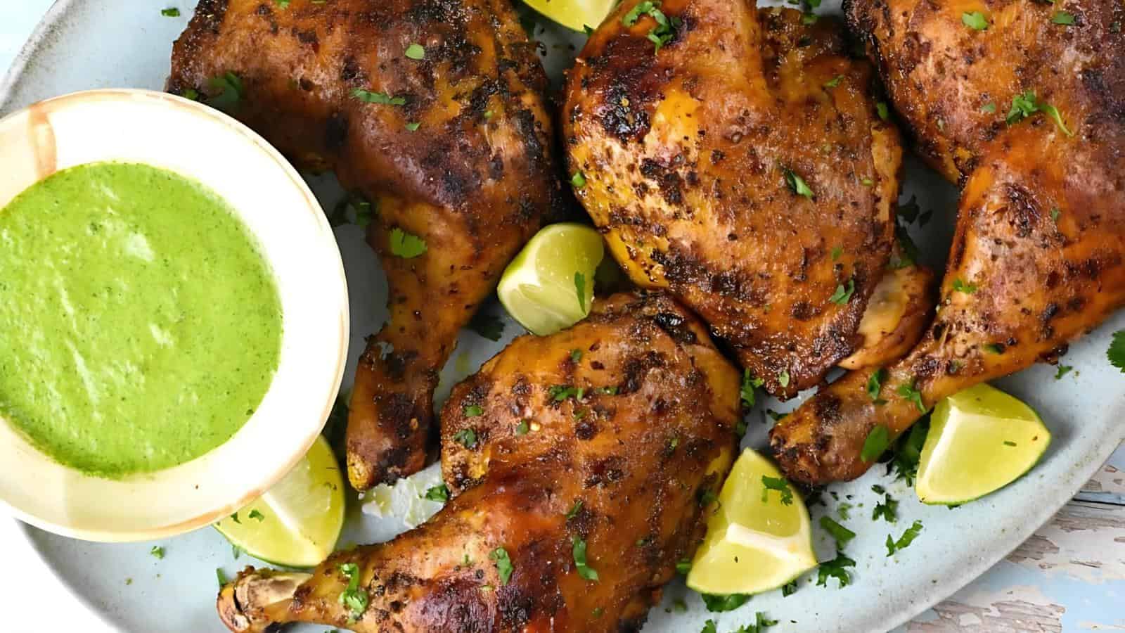 Grilled chicken legs seasoned with herbs, surrounded by lime wedges, and accompanied by a bowl of green sauce.