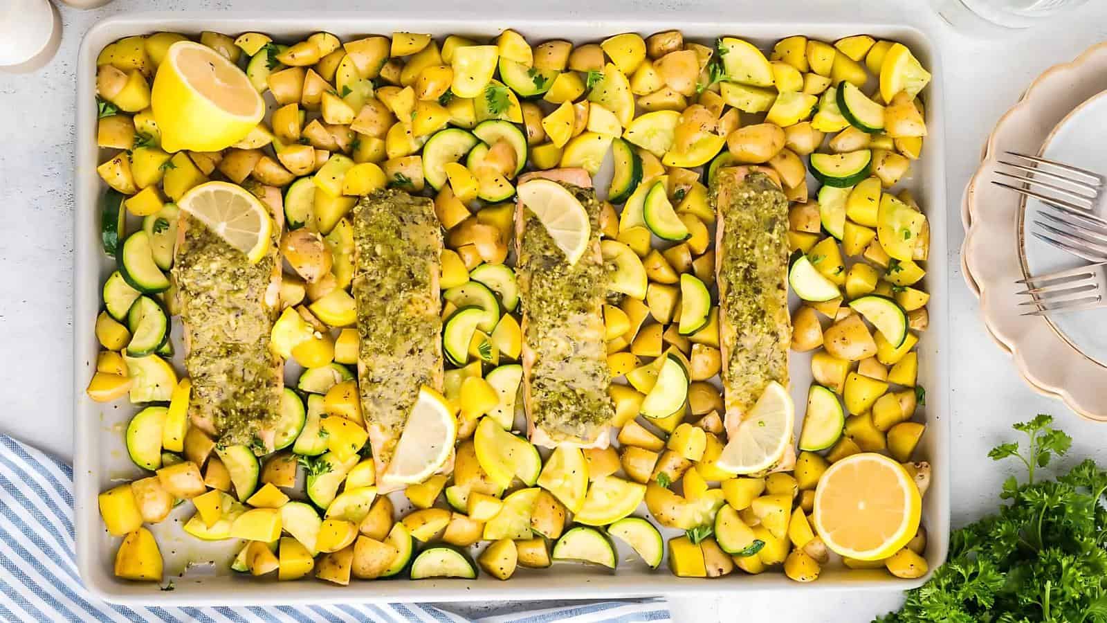 Baked fish fillets with lemon slices and herbs on a tray, surrounded by diced yellow squash, zucchini, and potatoes.