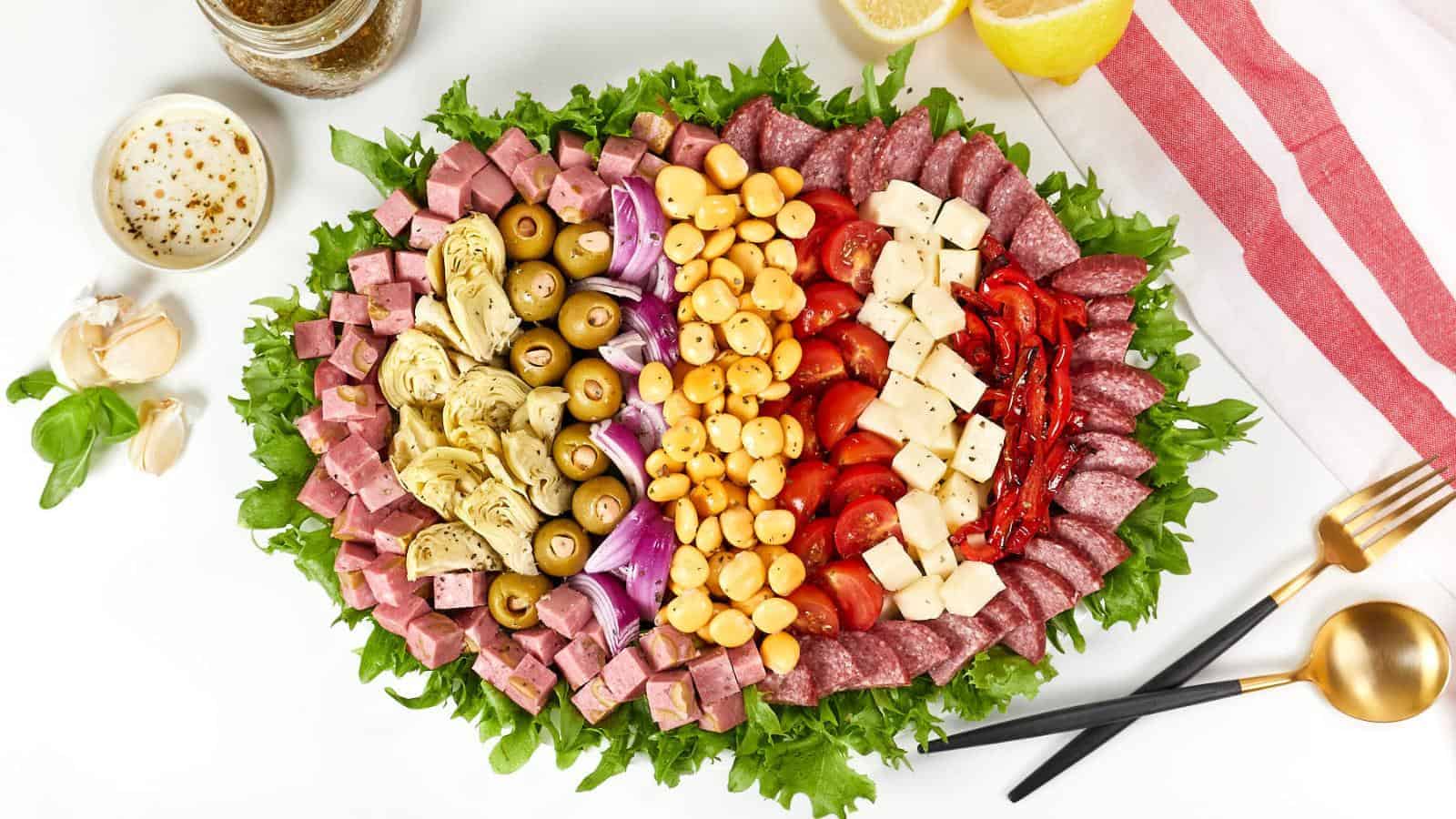 An oval antipasto platter with mixed meats, olives, artichokes, peppers, tomatoes, cheese, chickpeas, and leafy greens. Forks and a napkin are on the right, with dressing and garlic on the side.