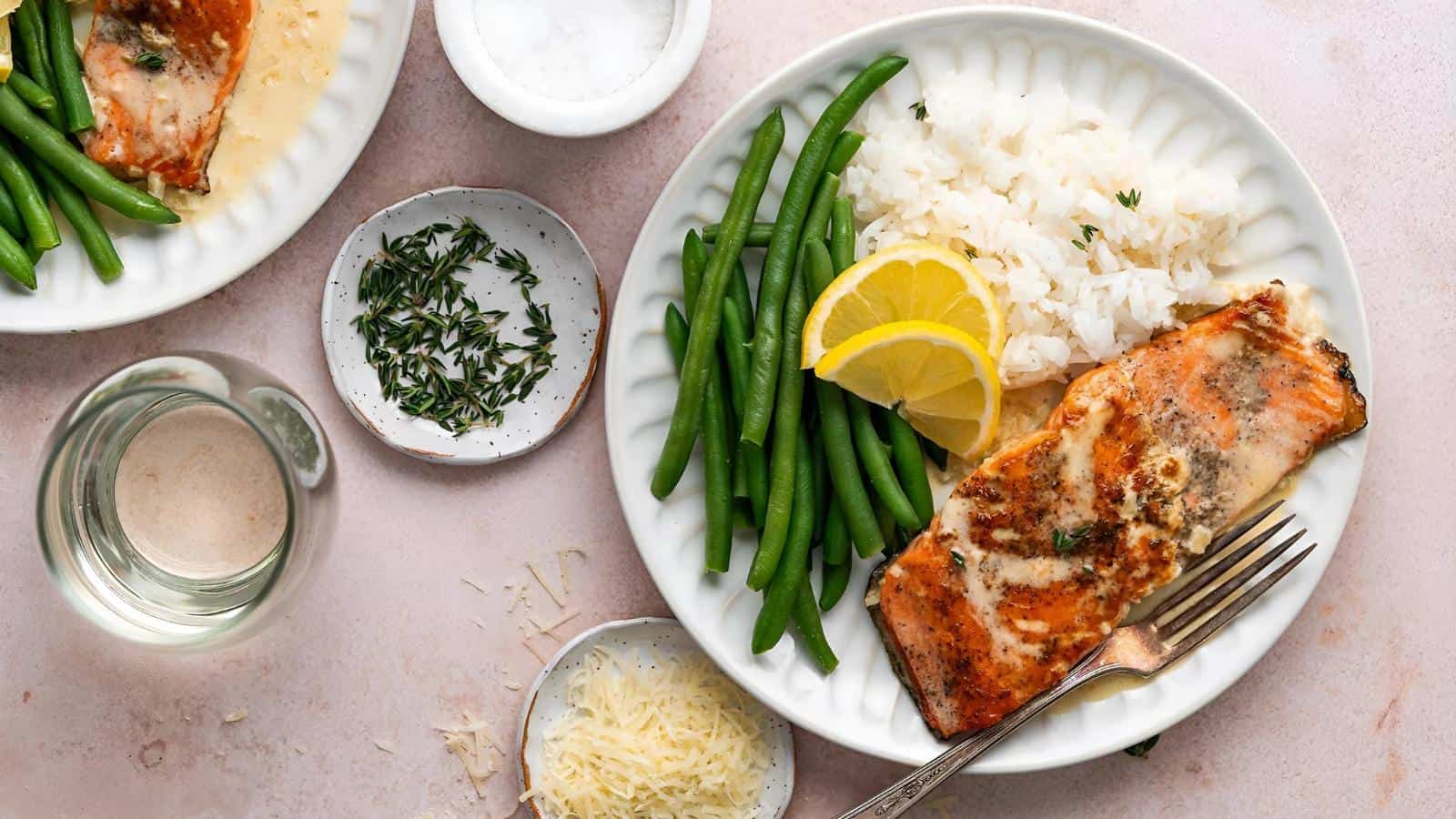 A plate with grilled salmon, green beans, white rice, and lemon slices. Nearby are a glass of white wine, grated cheese, herbs, and a small bowl of salt.