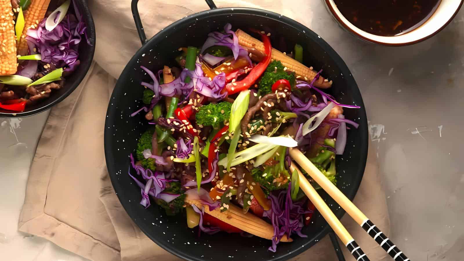 A bowl of stir-fried vegetables including broccoli, baby corn, red peppers, purple cabbage, and green onions, garnished with sesame seeds, next to a bowl of sauce and chopsticks.