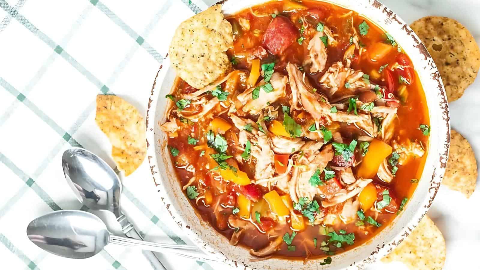 A bowl of chicken taco soup with shredded chicken, bell peppers, and chopped herbs. Surrounded by tortilla chips, with two spoons on a checkered cloth.