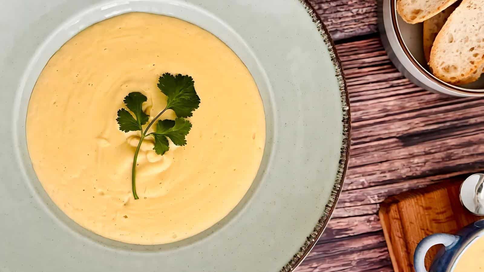 A bowl of creamy yellow soup garnished with a cilantro sprig, next to a container of sliced bread on a wooden surface.