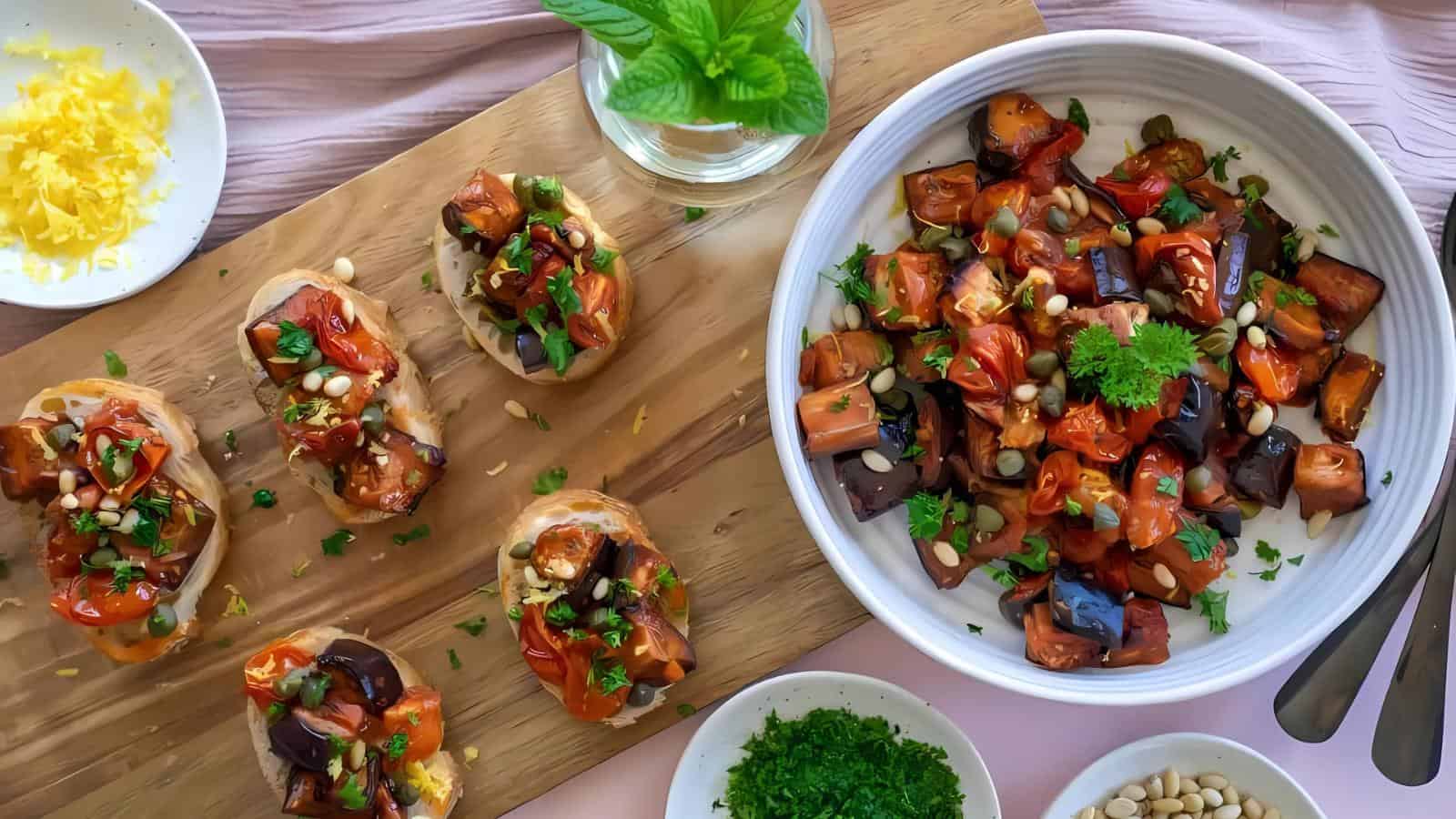 A white bowl and a wooden board with bruschetta topped with roasted vegetables, pine nuts, and herbs. Nearby are small bowls of shredded cheese, chopped parsley, and pine nuts.