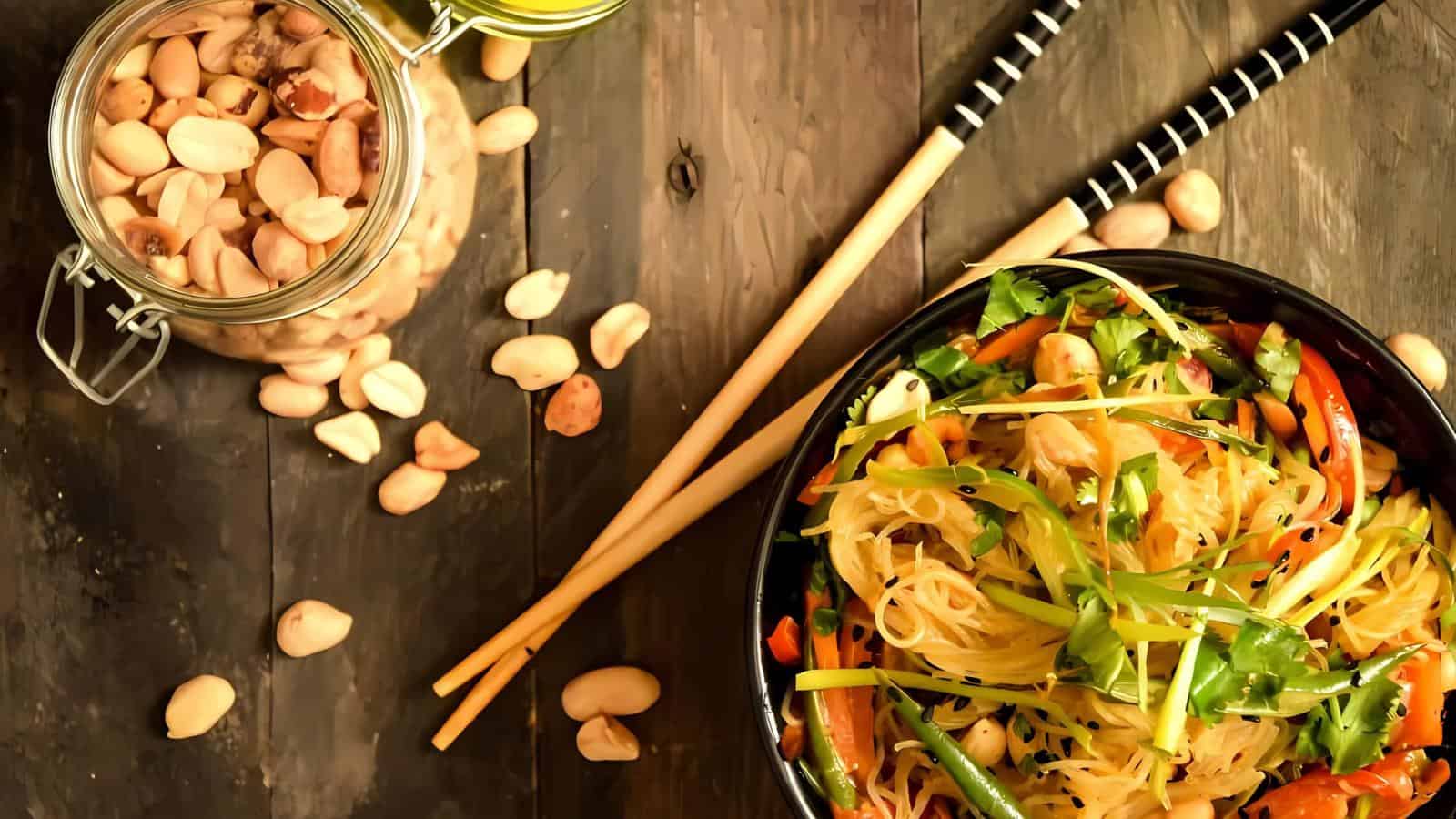 A bowl of noodle salad with vegetables and peanuts on a wooden surface. Chopsticks rest beside the bowl. A jar of peanuts is open nearby.