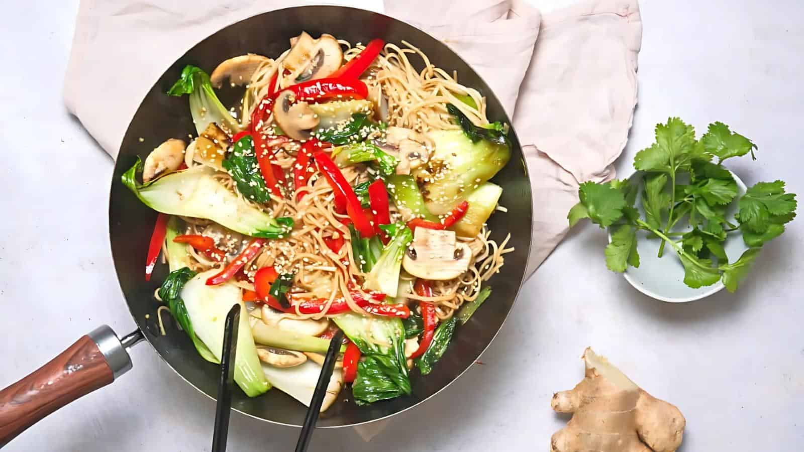 Stir-fried noodles with bok choy, red peppers, mushrooms, and sesame seeds in a wok. A bowl of cilantro and a piece of ginger are beside it.