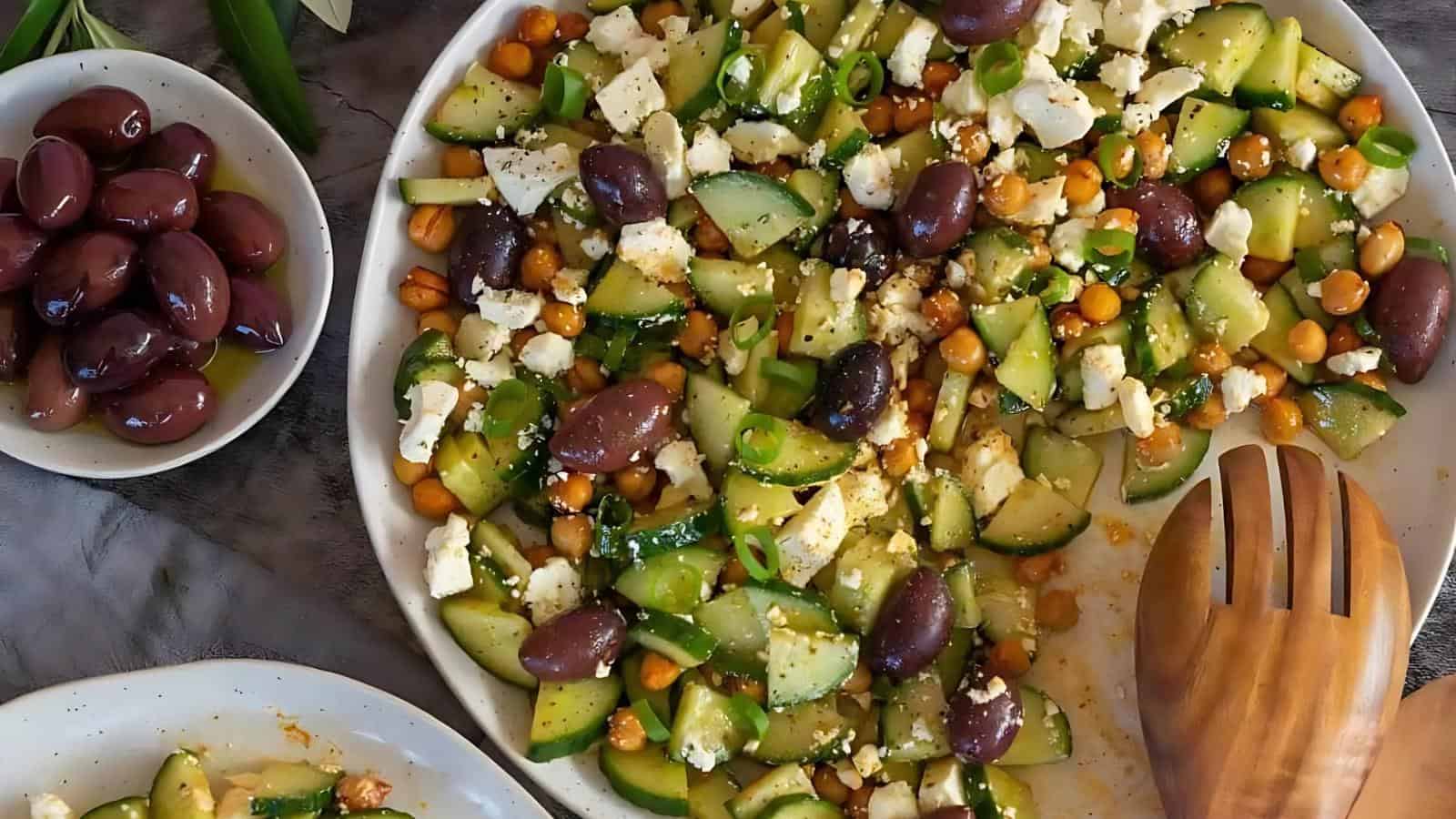 A plate of cucumber salad with feta, olives, chickpeas, and herbs, accompanied by a bowl of olives and a wooden spoon.