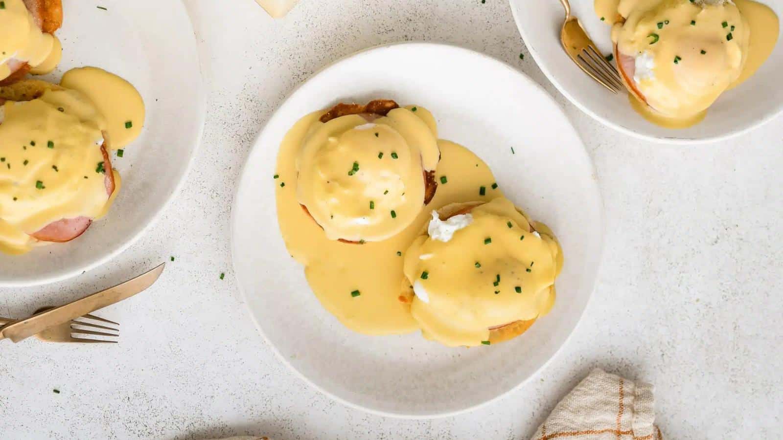 Plates of eggs Benedict topped with Hollandaise sauce and garnished with chives are on a white table, accompanied by cutlery and a napkin.