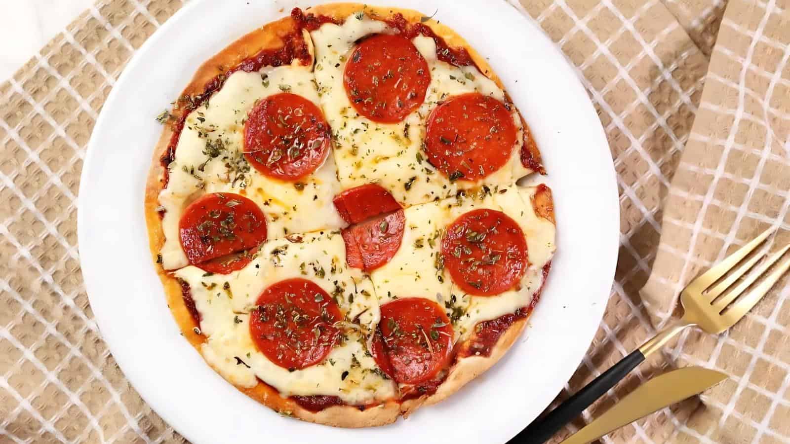 A pepperoni pizza on a white plate, topped with slices of pepperoni and melted cheese, garnished with herbs. A fork and knife are placed beside the plate on a checkered cloth.