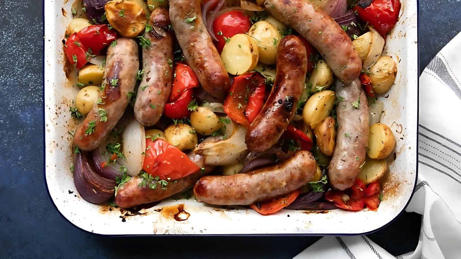 Baked sausages with potatoes, tomatoes, and onions in a white dish, garnished with herbs on a blue background.