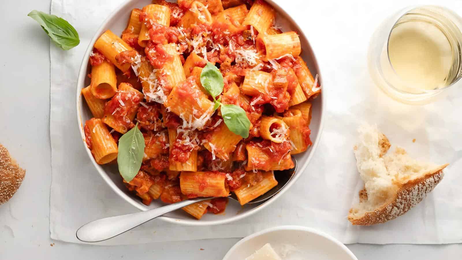 A bowl of rigatoni pasta with tomato sauce, garnished with basil and cheese. A glass of white wine and a piece of bread are on the side.
