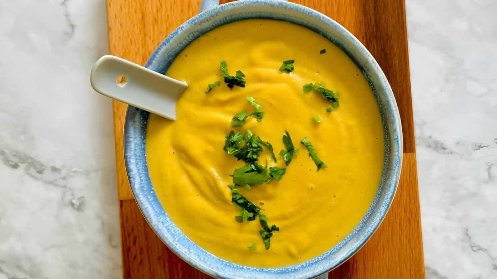 A bowl of creamy yellow soup garnished with chopped herbs, placed on a wooden surface.
