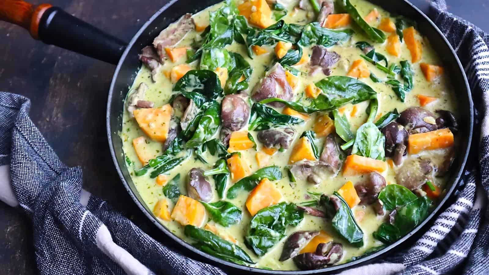 A skillet filled with a creamy curry includes spinach, sweet potatoes, mushrooms, and pieces of meat. A dark cloth is wrapped around the handle.