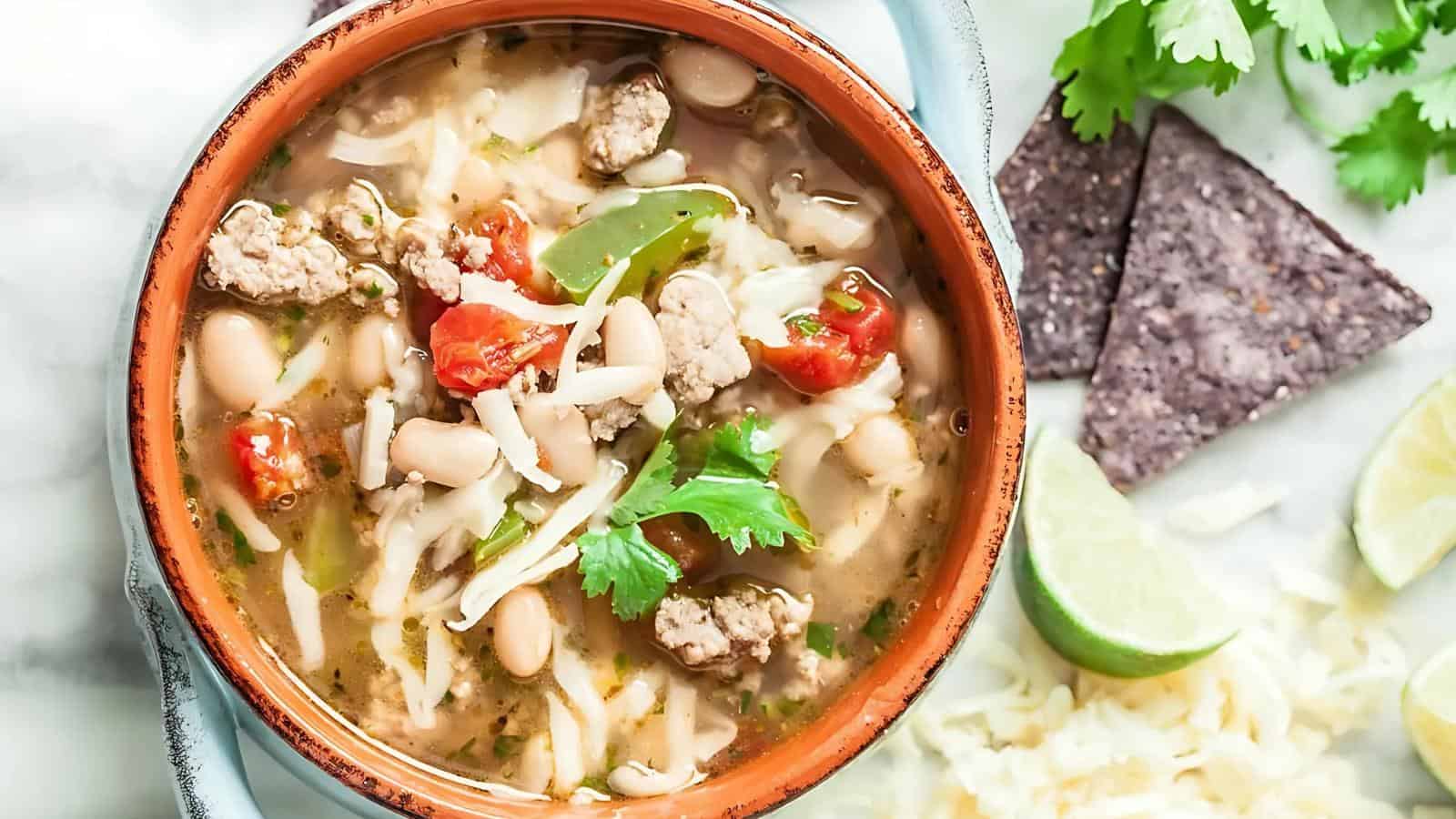 A bowl of soup with beans, meat, tomatoes, cheese, and cilantro. Blue corn tortilla chips, cheese, lime, and cilantro leaves are on the side.