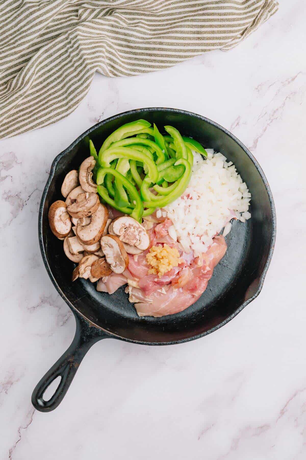 Chopped chicken, onions, green bell peppers, and mushrooms in a black skillet on a marble surface, with a striped cloth nearby.