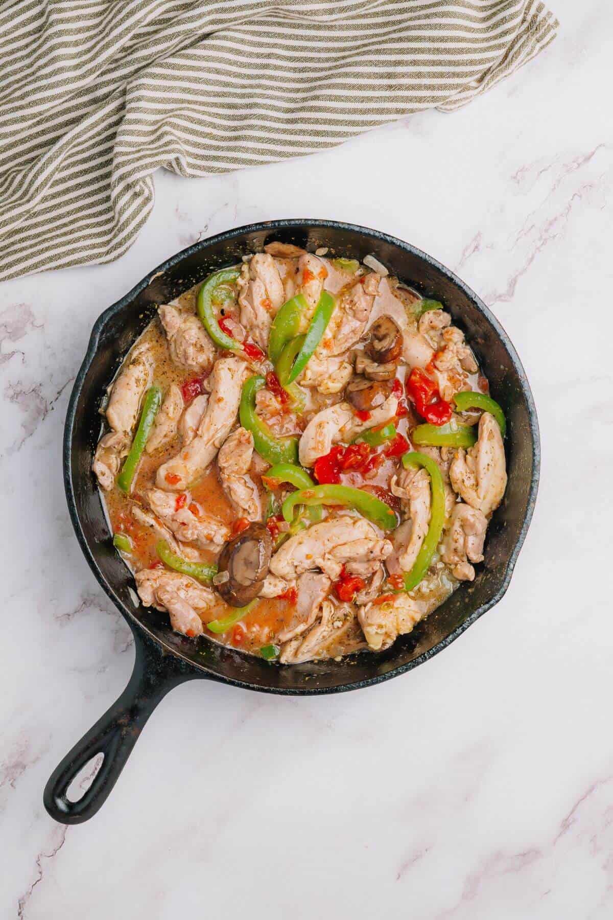 Skillet with cooked chicken strips, sliced green bell peppers, mushrooms, and diced red peppers in sauce, placed on a marble surface with a striped cloth nearby.