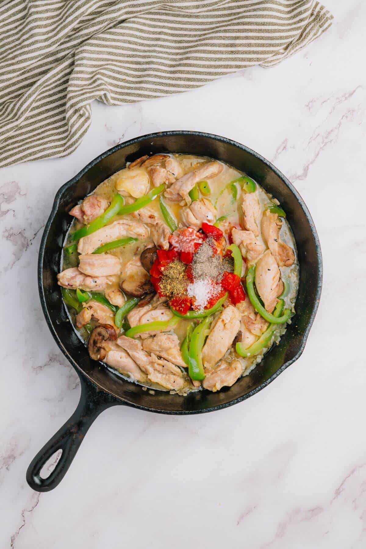 A skillet with chicken, green bell peppers, and red seasoning on a marble surface. A striped cloth is beside the skillet.