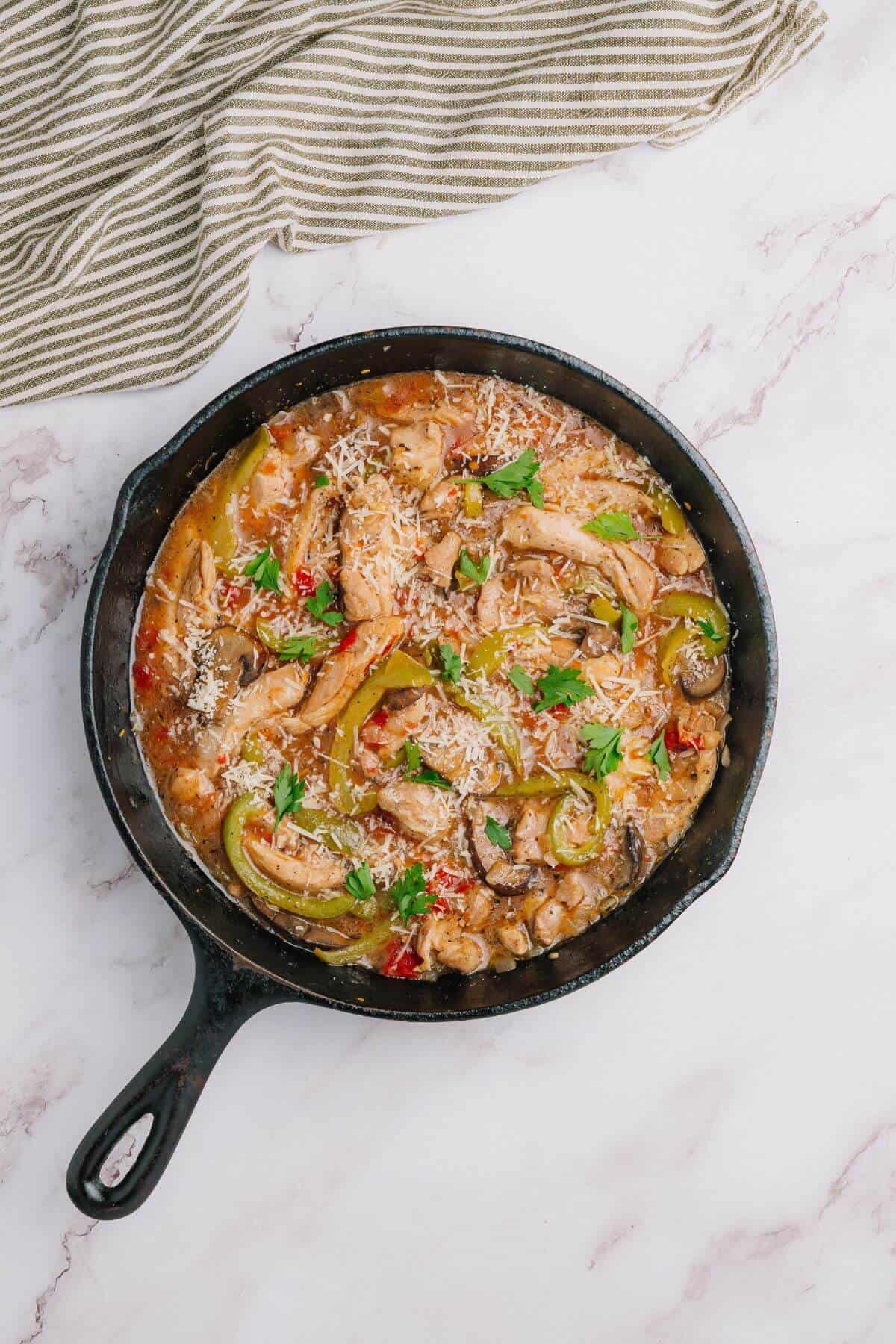 Skillet with chicken, bell peppers, mushrooms, and herbs in a creamy sauce, topped with grated cheese, on a marble surface.