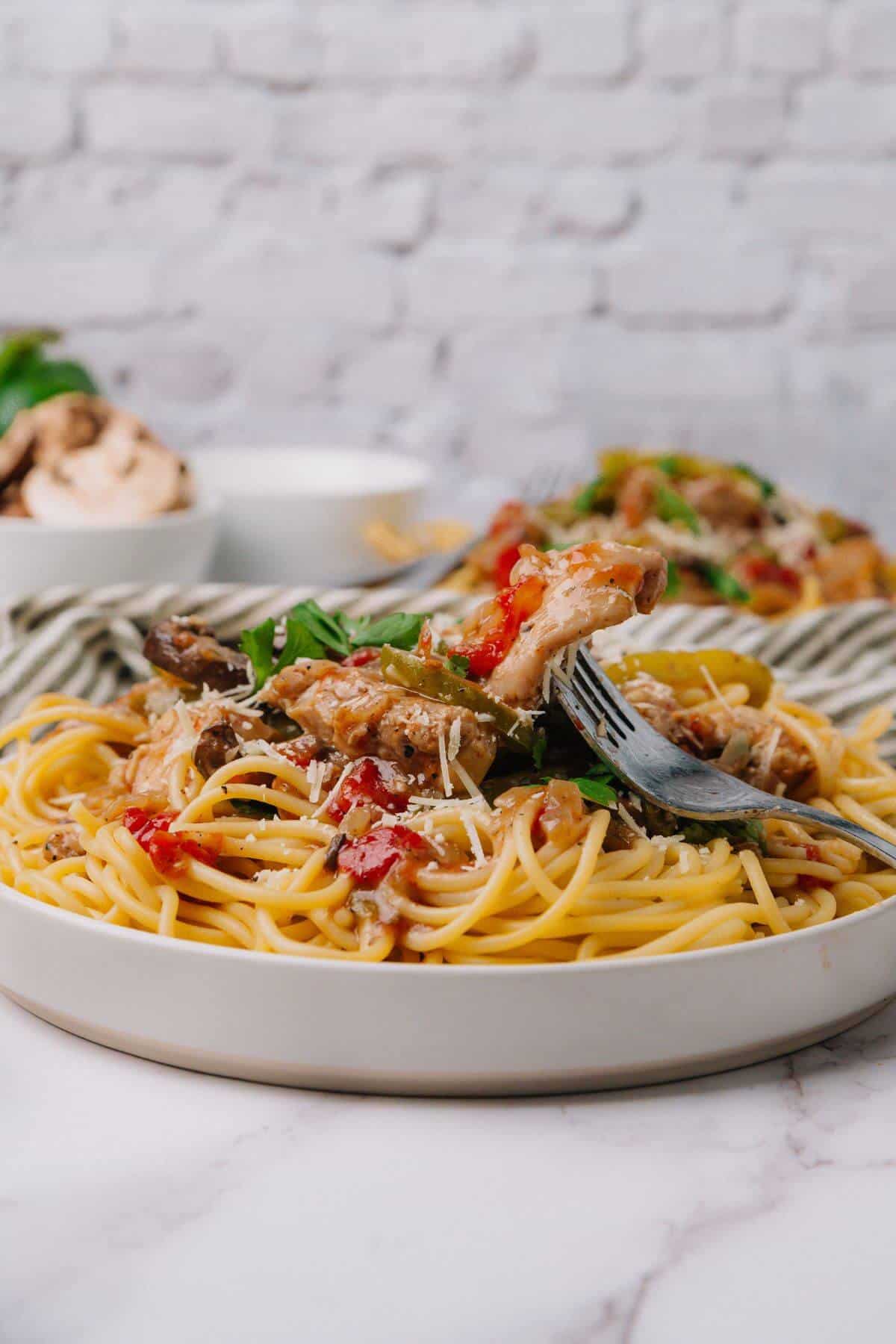 Plate of spaghetti topped with mushrooms, tomatoes, herbs, and grated cheese, with a fork twirling pasta, set on a table with striped cloth and a bowl in the background.
