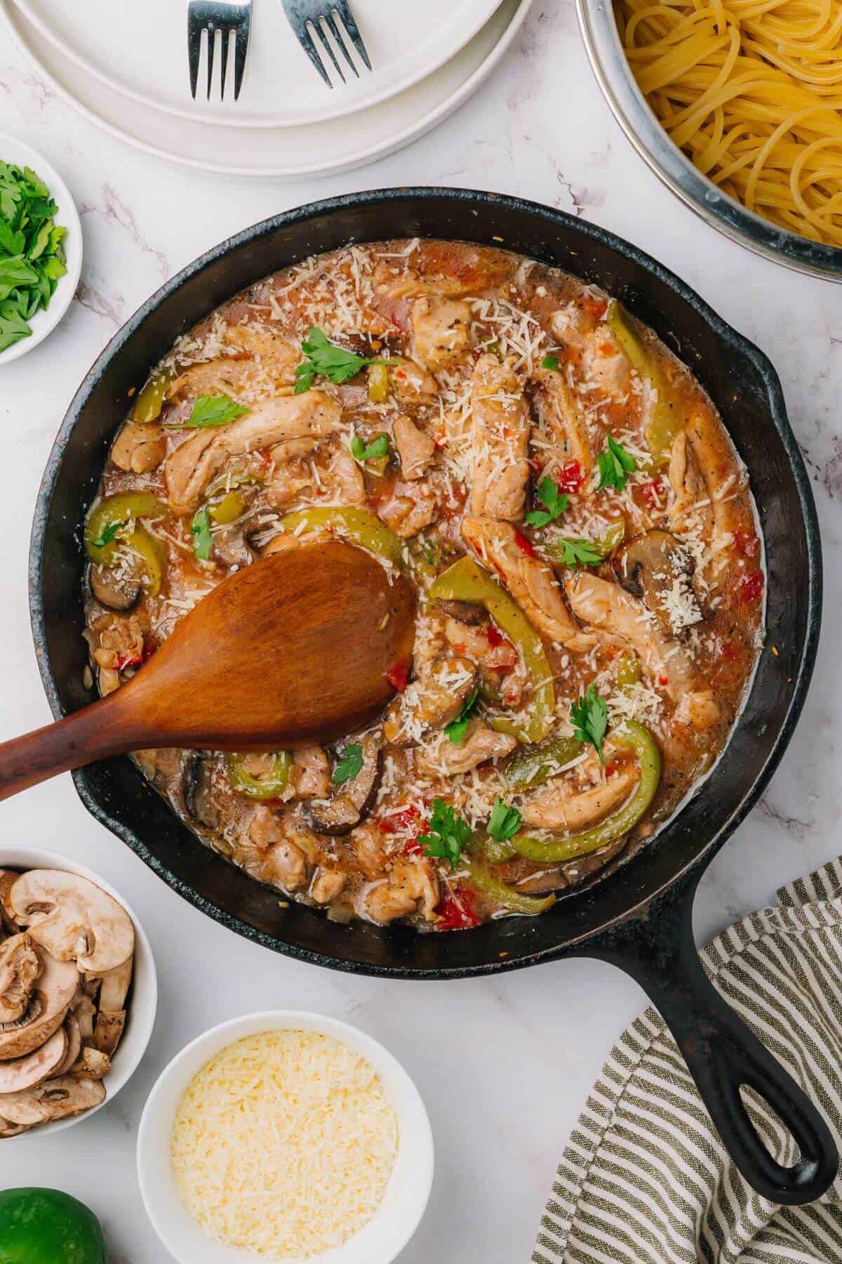 A skillet with cooked chicken strips, green bell peppers, mushrooms, and sauce, topped with grated cheese and parsley. Nearby, uncooked pasta and bowls of sliced mushrooms and grated cheese.