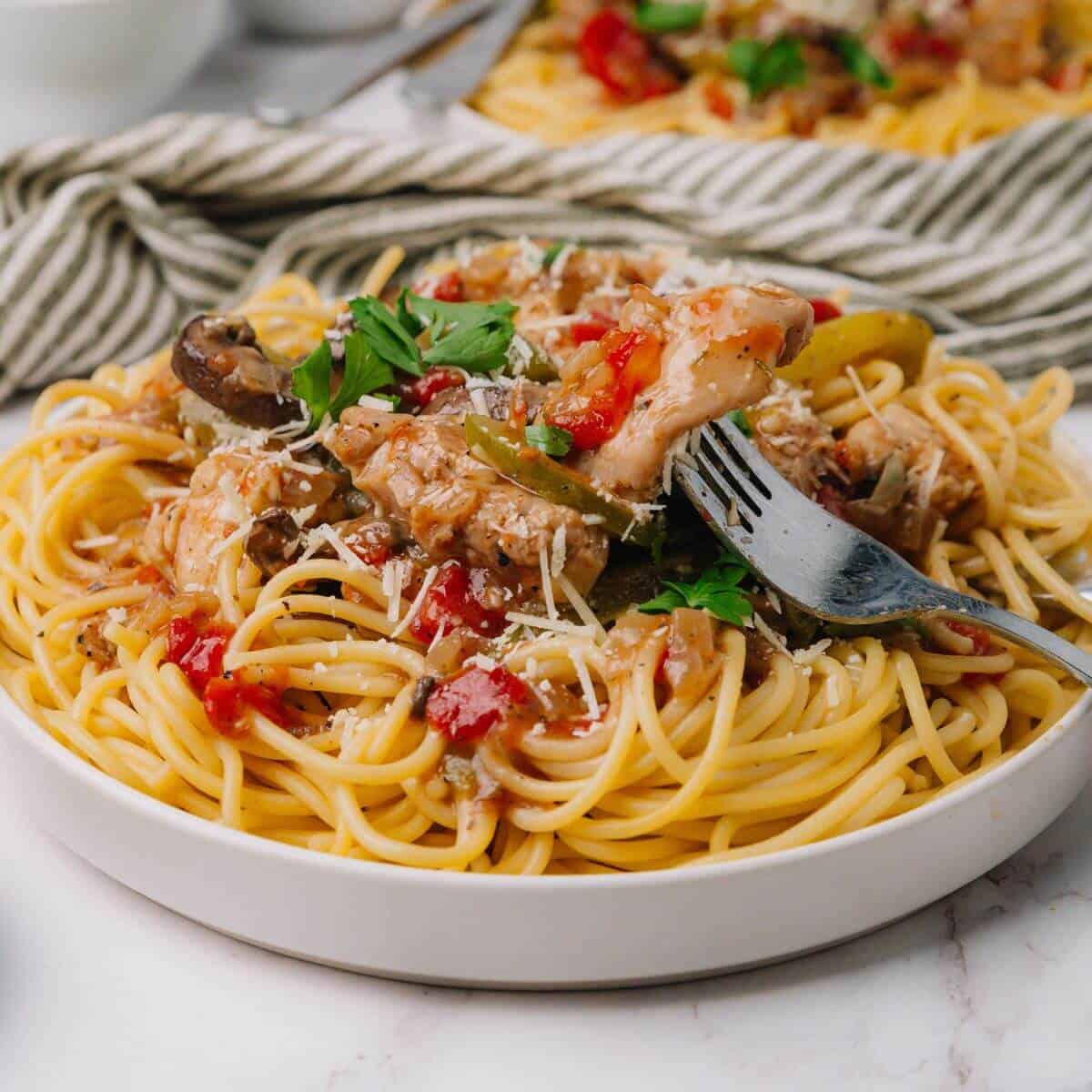 Plate of spaghetti with tomato sauce, chicken, and vegetables. A fork is twirling some pasta. Garnished with herbs. A striped napkin is in the background.